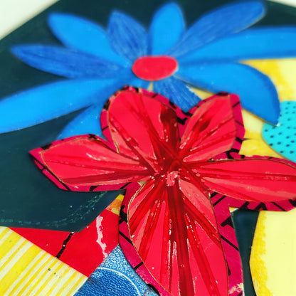 A close-up of a colorful abstract floral artwork with layered paper flowers in bright blue, red, yellow, and black.