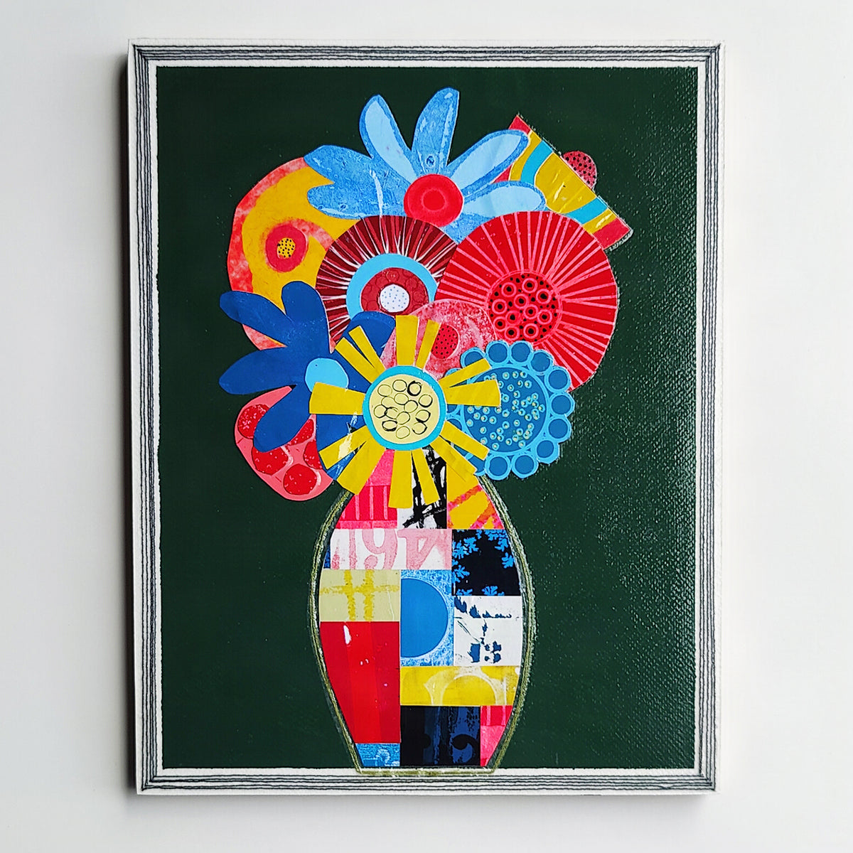 A framed abstract floral artwork hanging on a light wall. The piece features a dark green background with a central vase and layered flower-like shapes in bright blue, red, yellow, pink, and white, enclosed in a silver frame.