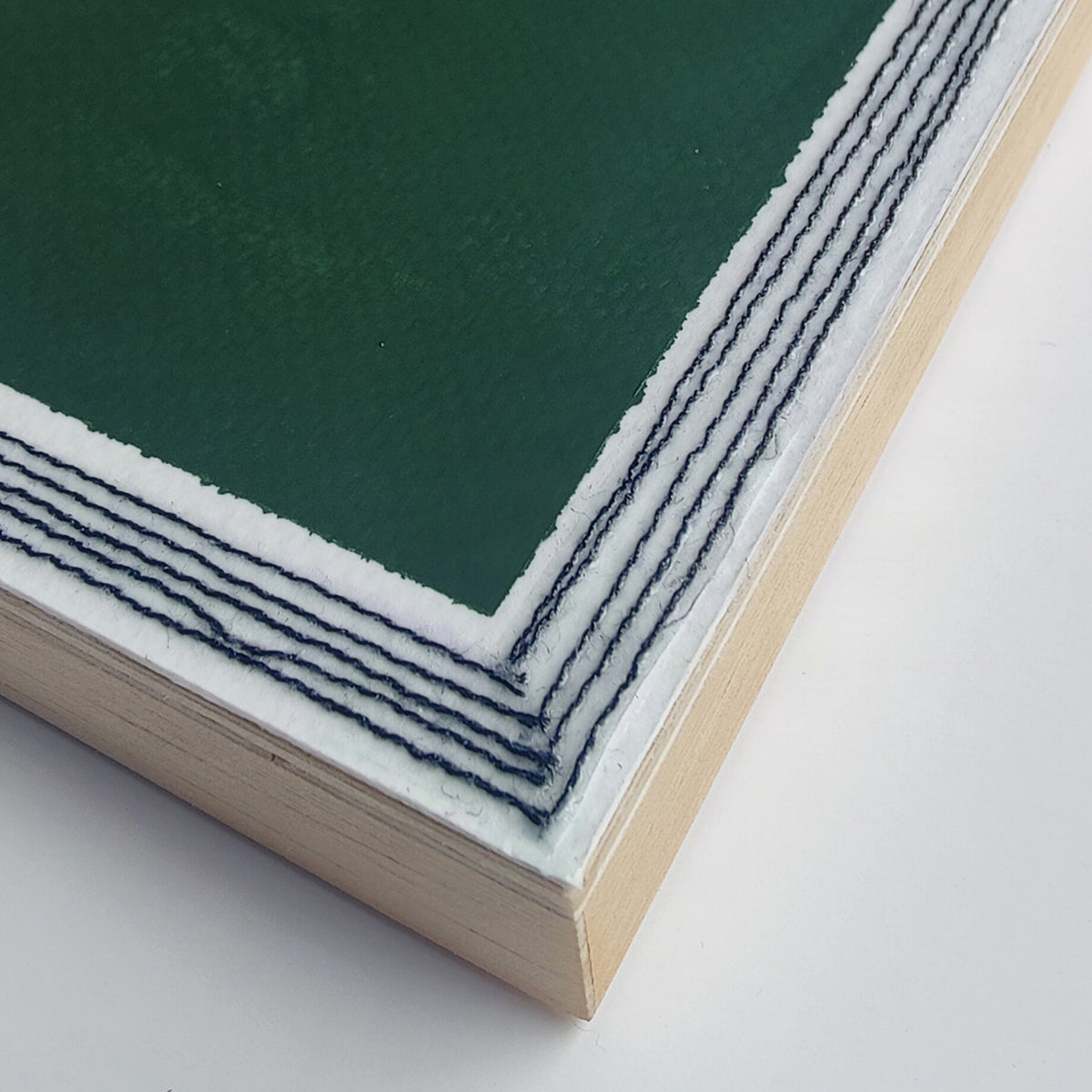 Close-up of a square art print or panel with a dark green center, white border with black machine stitching, and a light natural wood frame