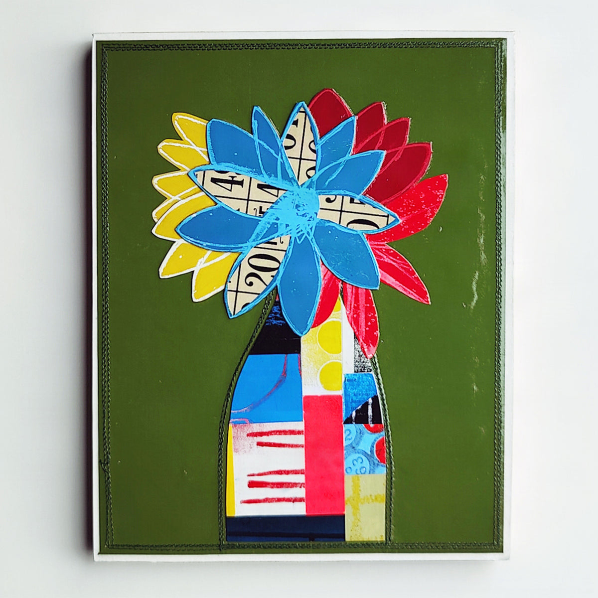 A framed abstract floral artwork on a white wall. The piece features layered flower shapes in blue, red, yellow, and white with black patterned centers, set against an olive green background and a dark green vase-like form below.