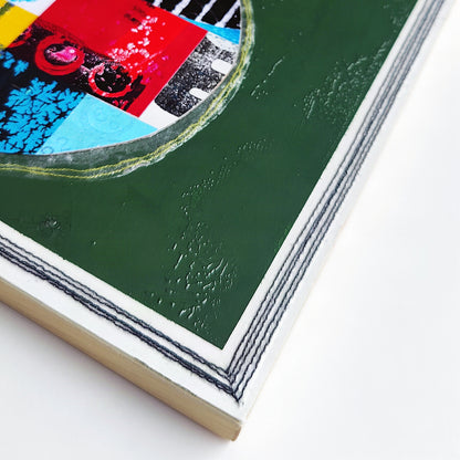 A close-up of a colorful mixed-media artwork on a square wooden panel, showing a dark green textured background, a circular collage with red, blue, black, and white elements, and a black stitched border.