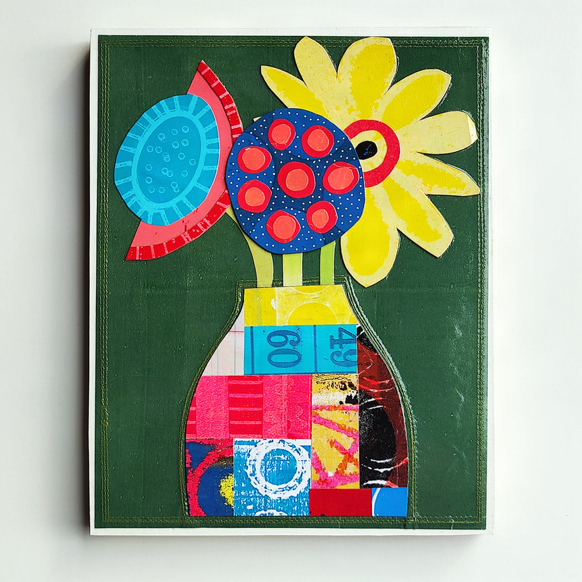A colorful abstract floral artwork on a dark green background, featuring a yellow flower, blue and red circular motifs, and a patchwork vase in bright pink, blue, yellow, and red tones, photographed against a light neutral wall.