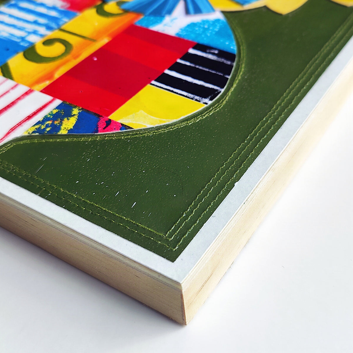 Close-up of a framed art print showing a colorful abstract collage design with a prominent olive green stitched border and light wood frame on a white background.